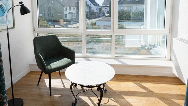 A room with a large window, a green chair, a white table, and a floor lamp. - Klusenhof (Lippstadt)