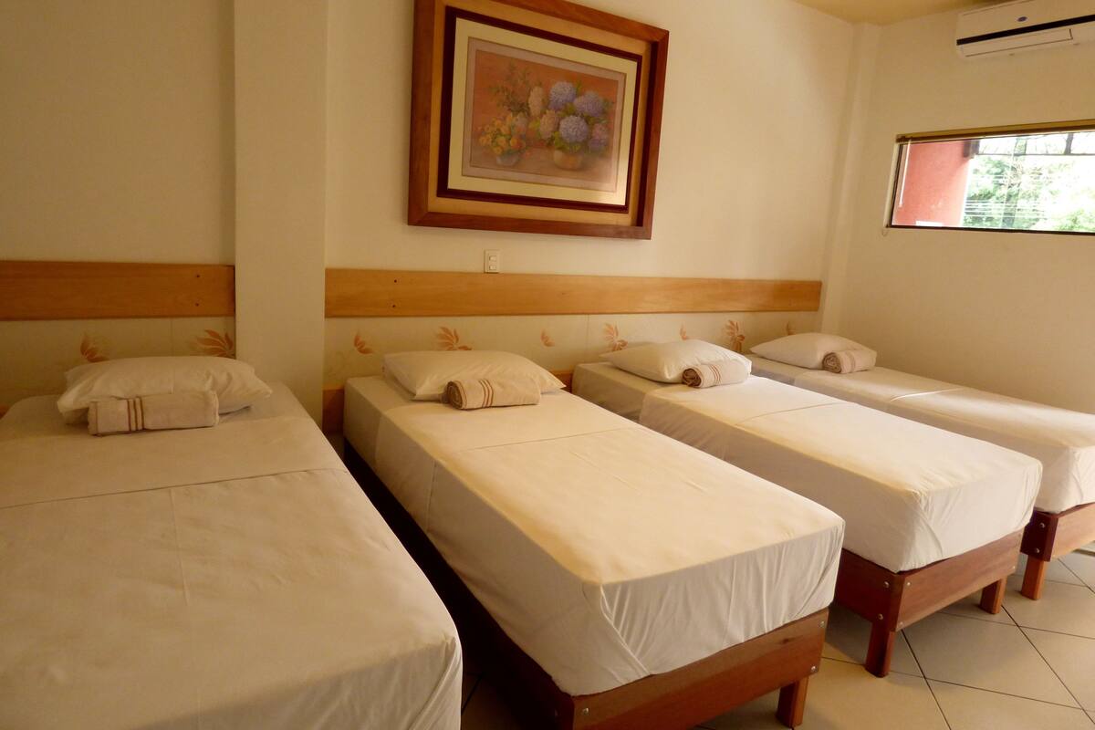 A hotel room with three single beds, each with a wooden headboard and white bedding. There is a framed picture of flowers on the wall above the beds.