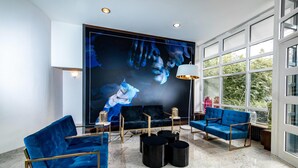 A modern lounge with blue sofas, a large abstract painting, and a view of greenery through large windows. - Aspire Elements Reutlingen, Trademark Collection by Wyndham (Reutlingen)