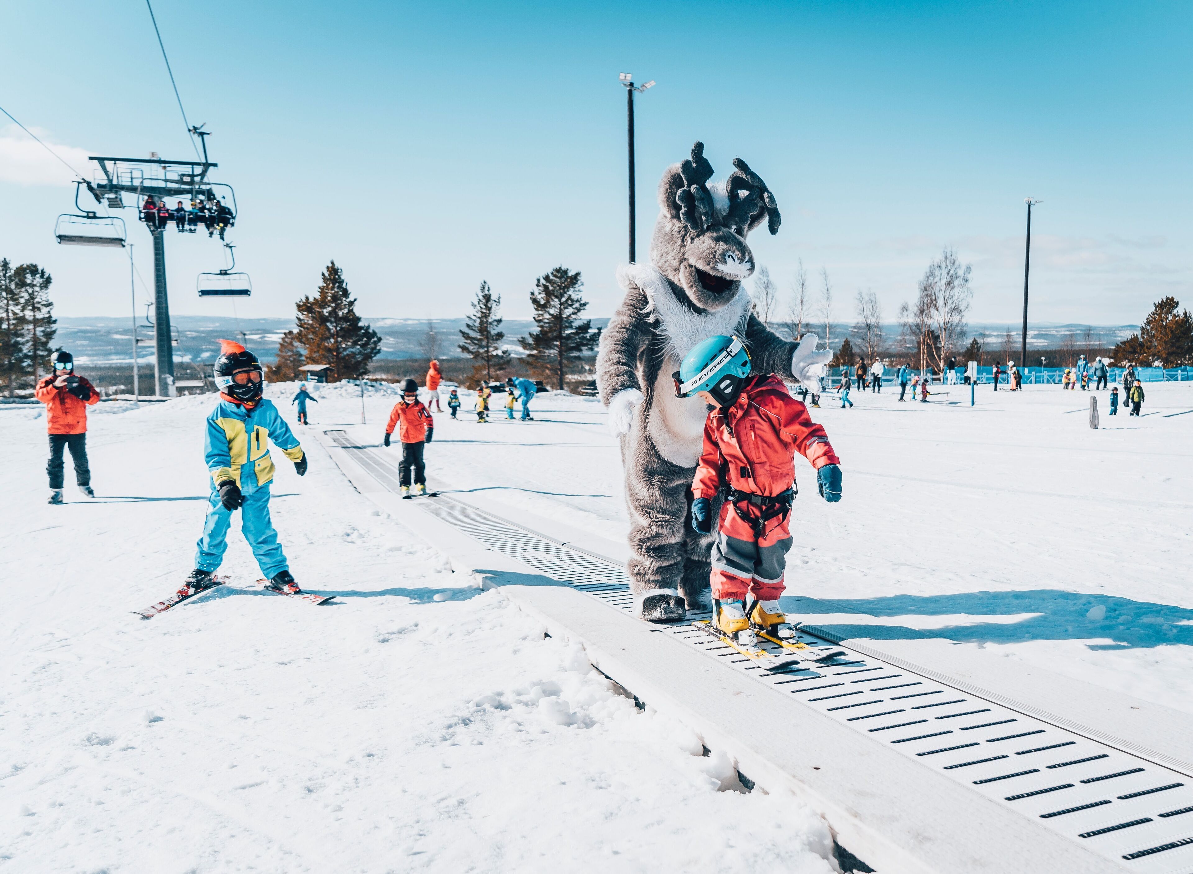 Une piste de ski avec un télésiège en arrière-plan, une mascotte et des skieurs.