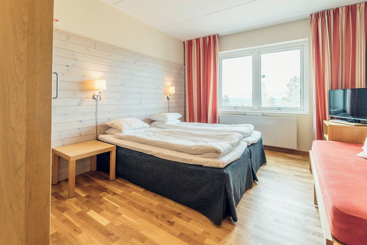 A modern hotel room with a wooden floor, a bed with a grey bedspread, a bedside table, a TV on a wooden stand, and a window with curtains.