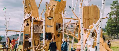 A wooden play structure with climbing nets and slides, surrounded by birch trees.