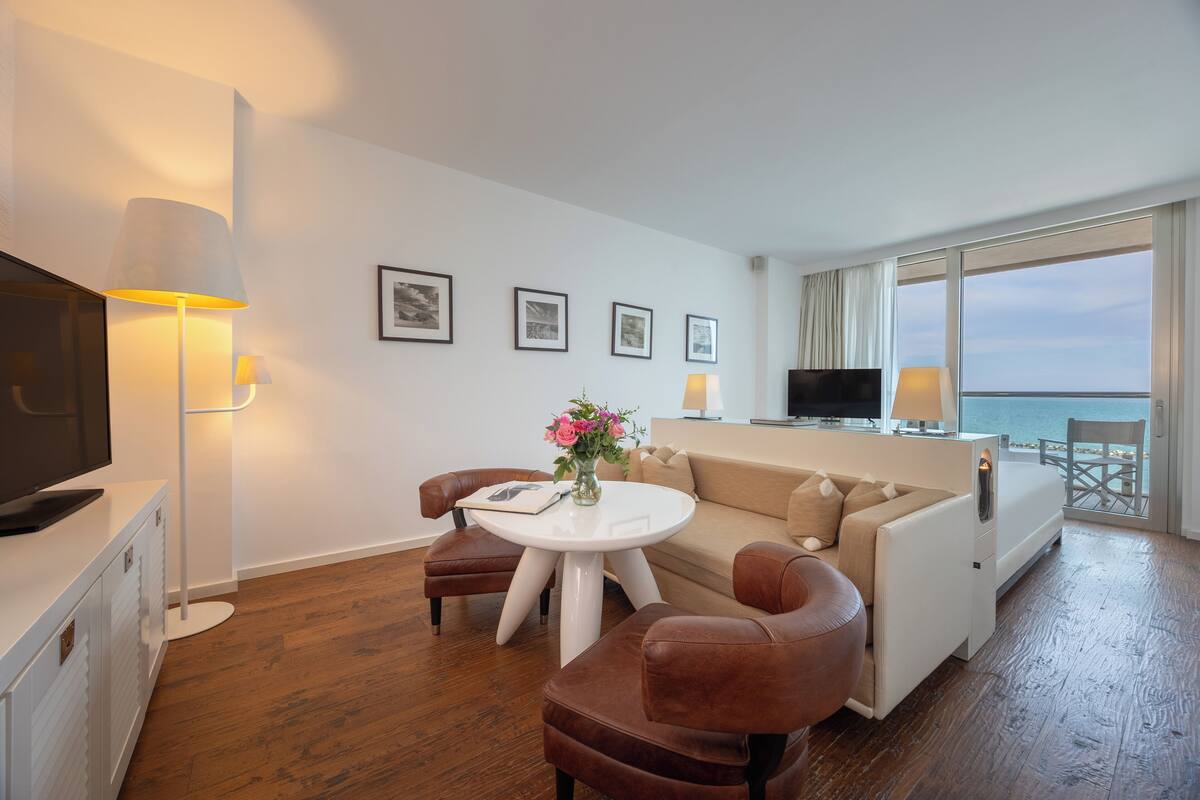 A modern hotel room with a wooden floor, a white sofa, a round coffee table with a vase of flowers, a flat-screen TV, and a balcony with a view of the sea.
