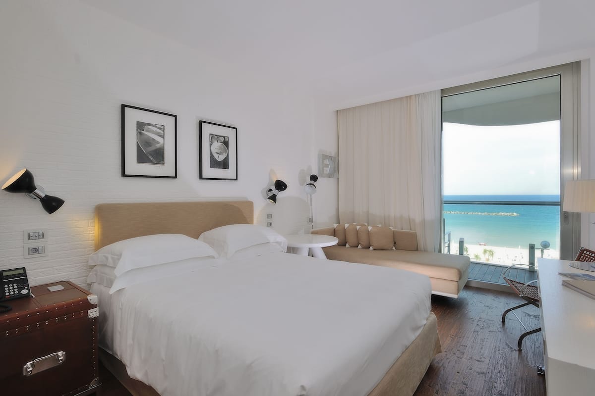 A hotel room with a bed, a sofa, a desk, a chair, a lamp, a phone, and a suitcase. There are two framed pictures on the wall and a view of the ocean through a large window.