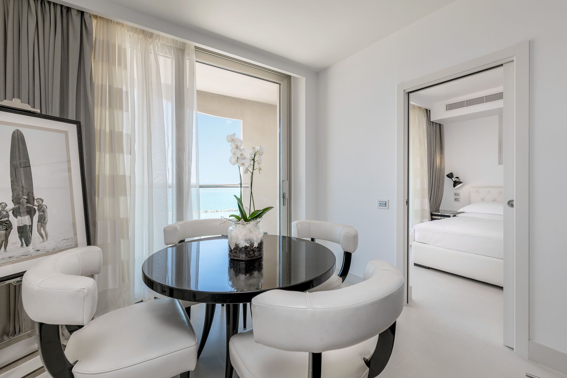 A modern hotel room with a round dining table, white leather chairs, a bed with white bedding, a large window with sheer curtains, and a framed picture on the wall.