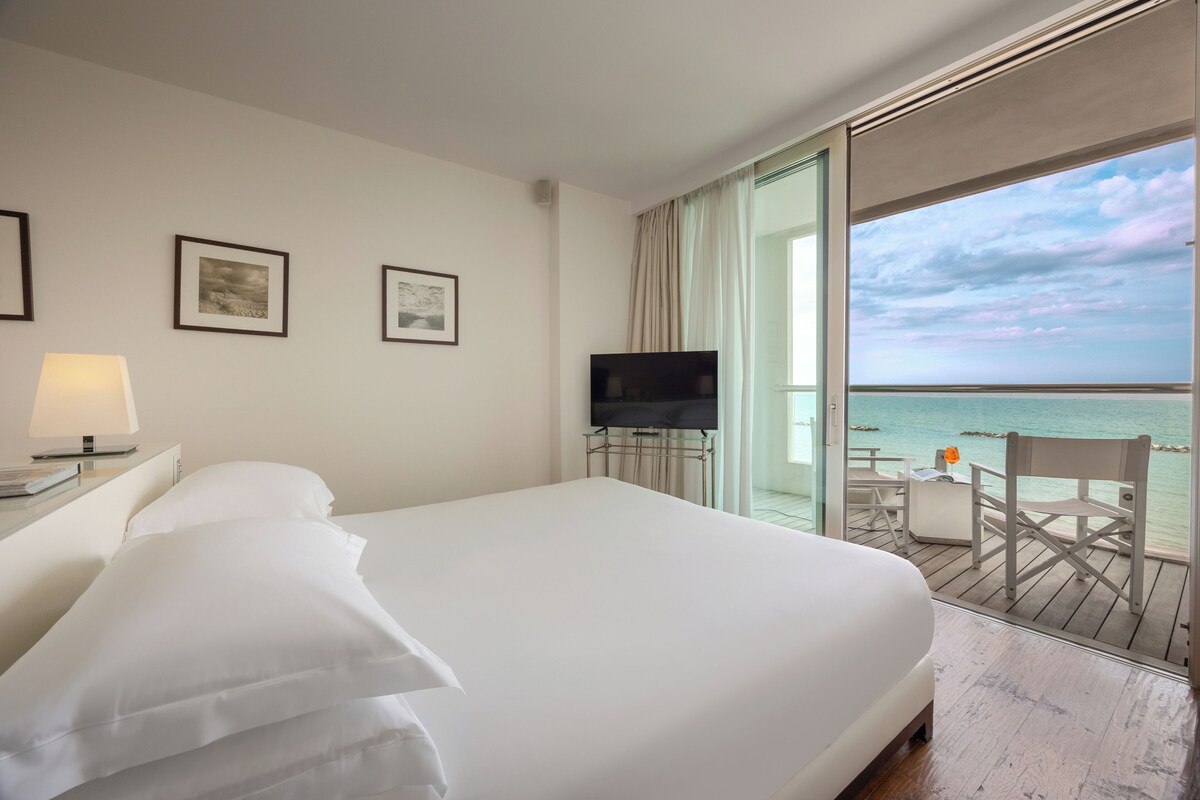 A hotel room with a large bed, a TV on a stand, a desk with a lamp, and a balcony with a view of the sea.