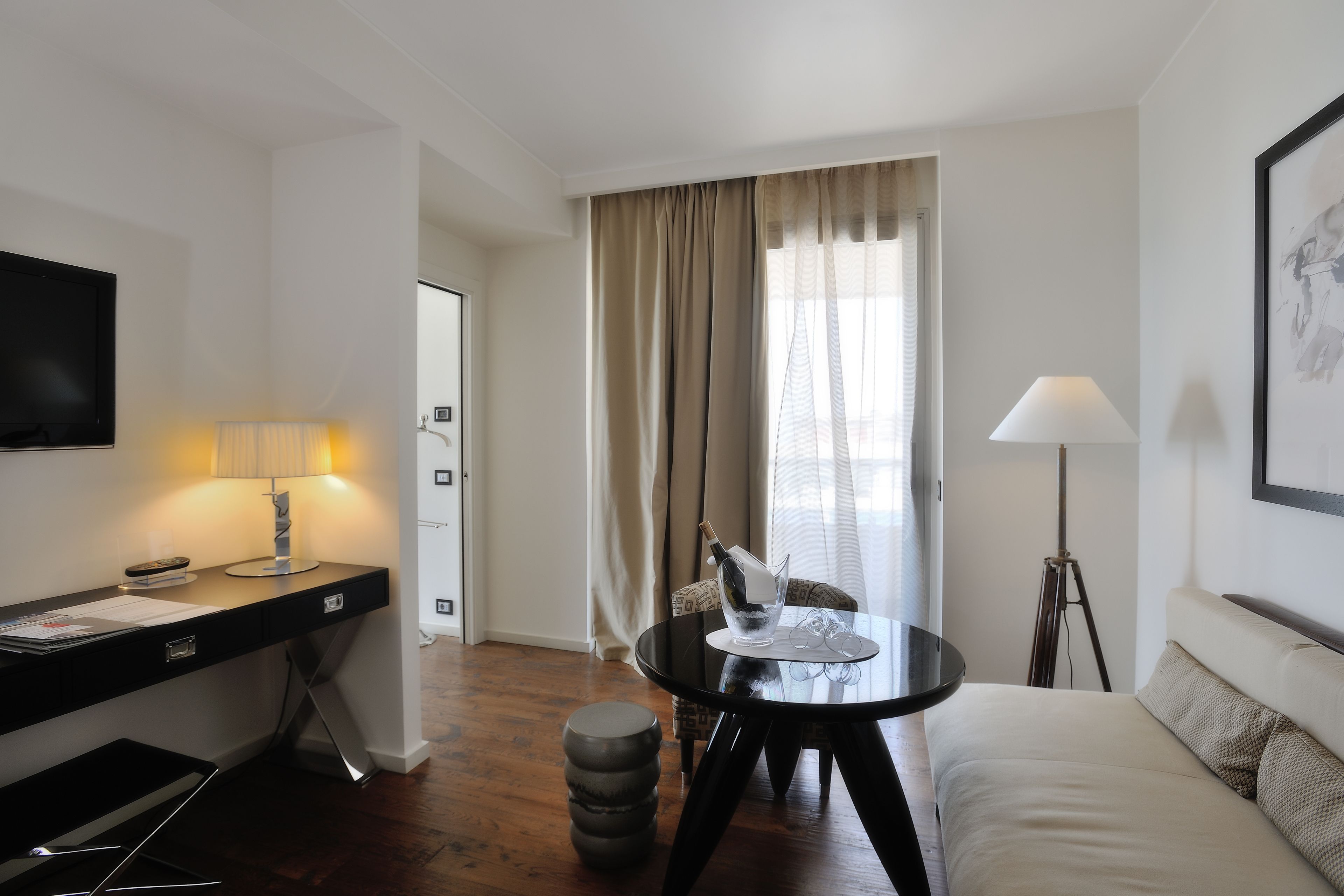 A hotel room with a bed, a desk with a lamp, a chair, a table with a bottle and glasses, and a floor lamp.