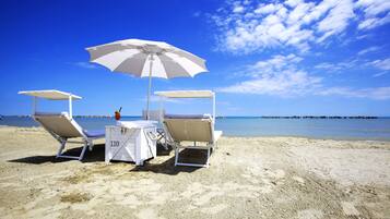 A beachside table with two chairs and a white umbrella, set on a sandy beach with a clear blue sky and calm sea in the background.