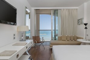 A hotel room with a bed, a desk with a laptop, a chair, a sofa, a small table, a TV, and a view of the sea through a large window.