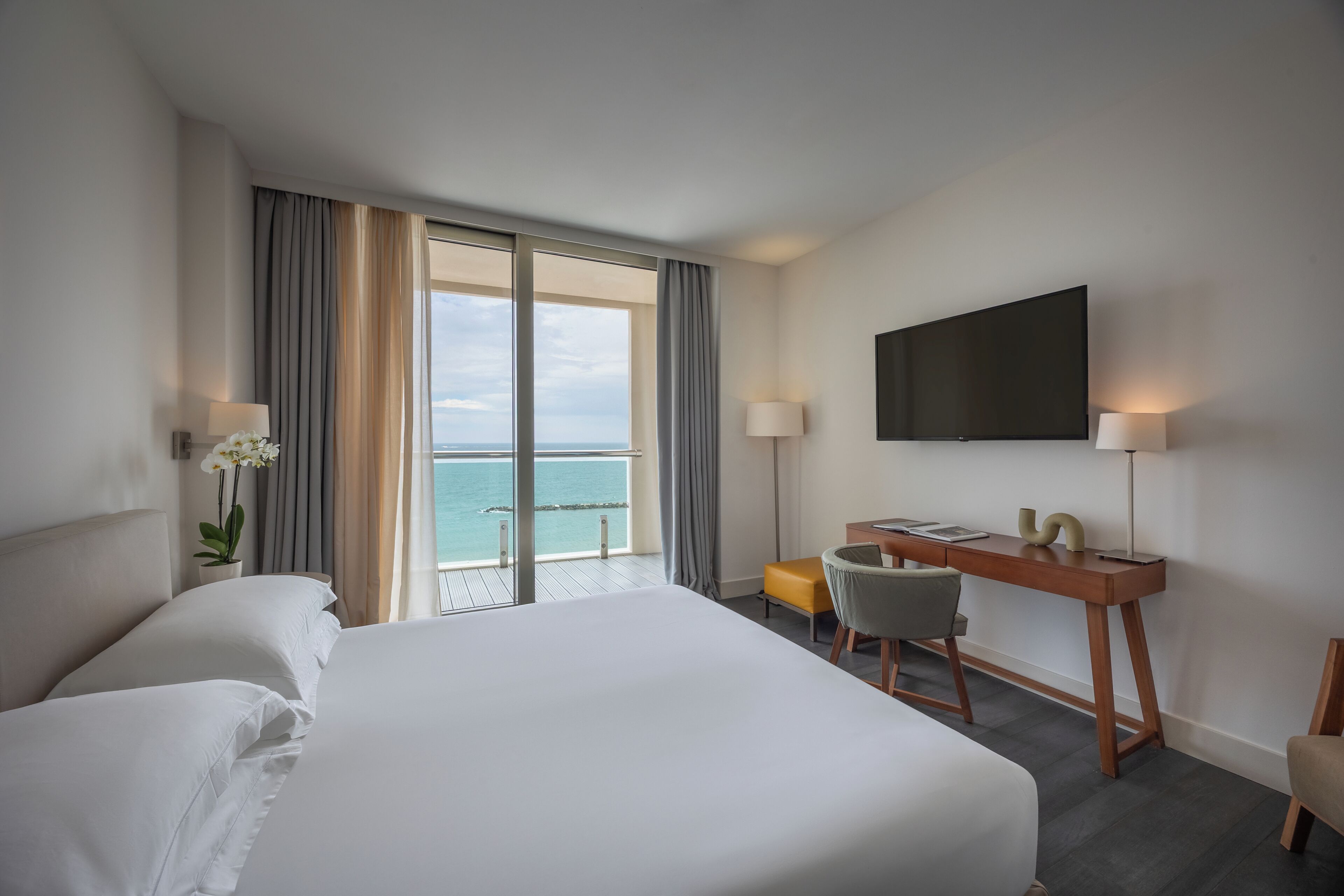 A hotel room with a large bed, a desk with a chair, a TV mounted on the wall, and a view of the sea through large windows.