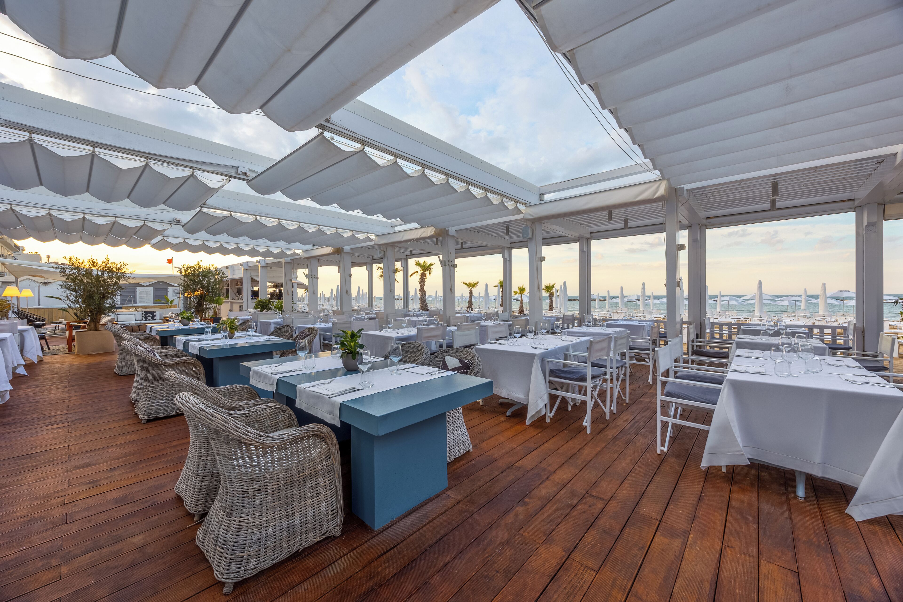 An outdoor dining area with white tables and wicker chairs, covered by a white canopy, overlooking a cityscape.