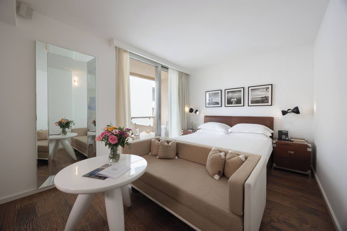 A hotel room with a bed, a sofa, a small table with a vase of flowers, a mirror, and a nightstand with a lamp.