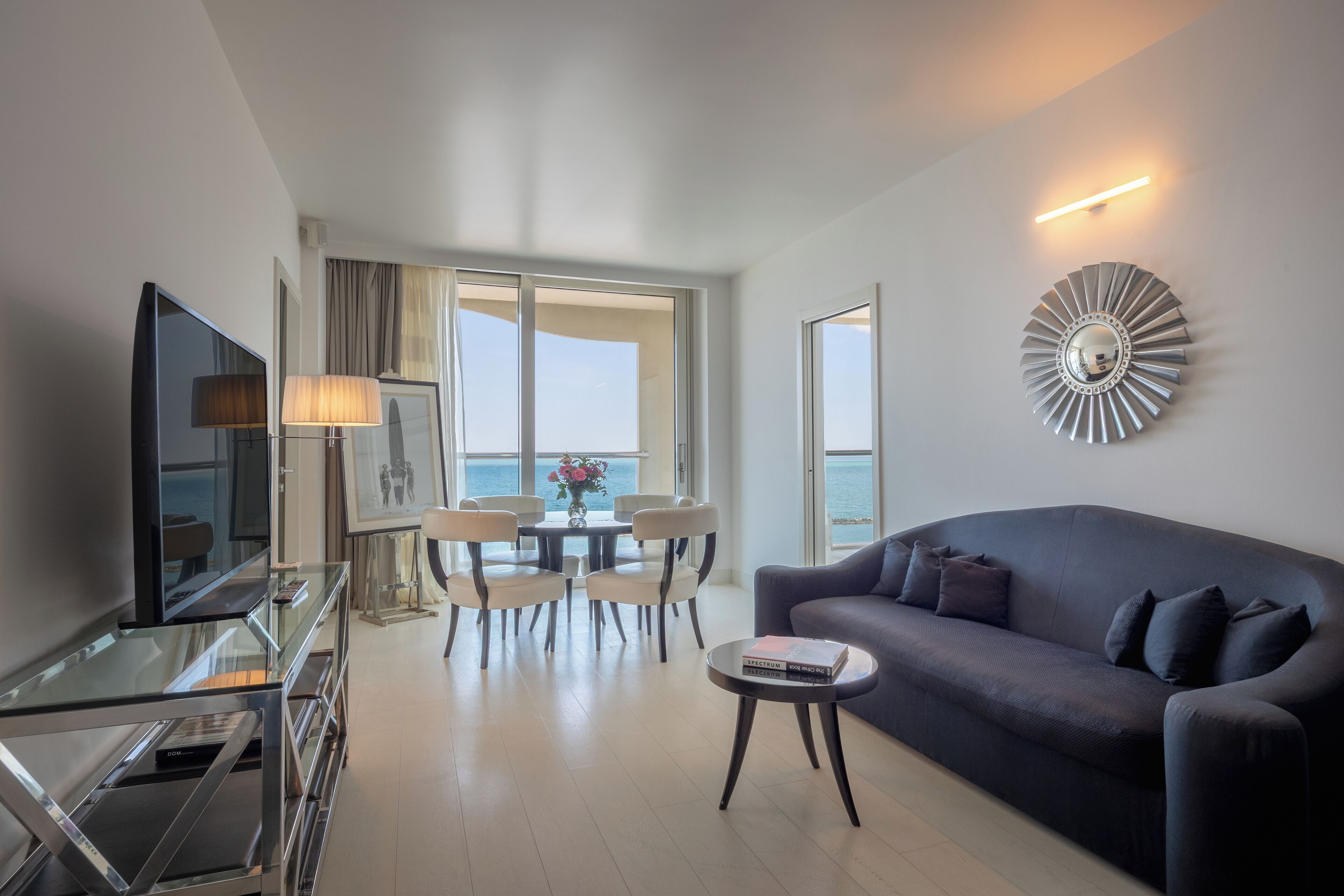 Een moderne woonkamer met een donkerblauwe bank, een glazen salontafel en een flatscreen-televisie. Er is een eethoek met een tafel en stoelen, en een groot raam met uitzicht op de zee.