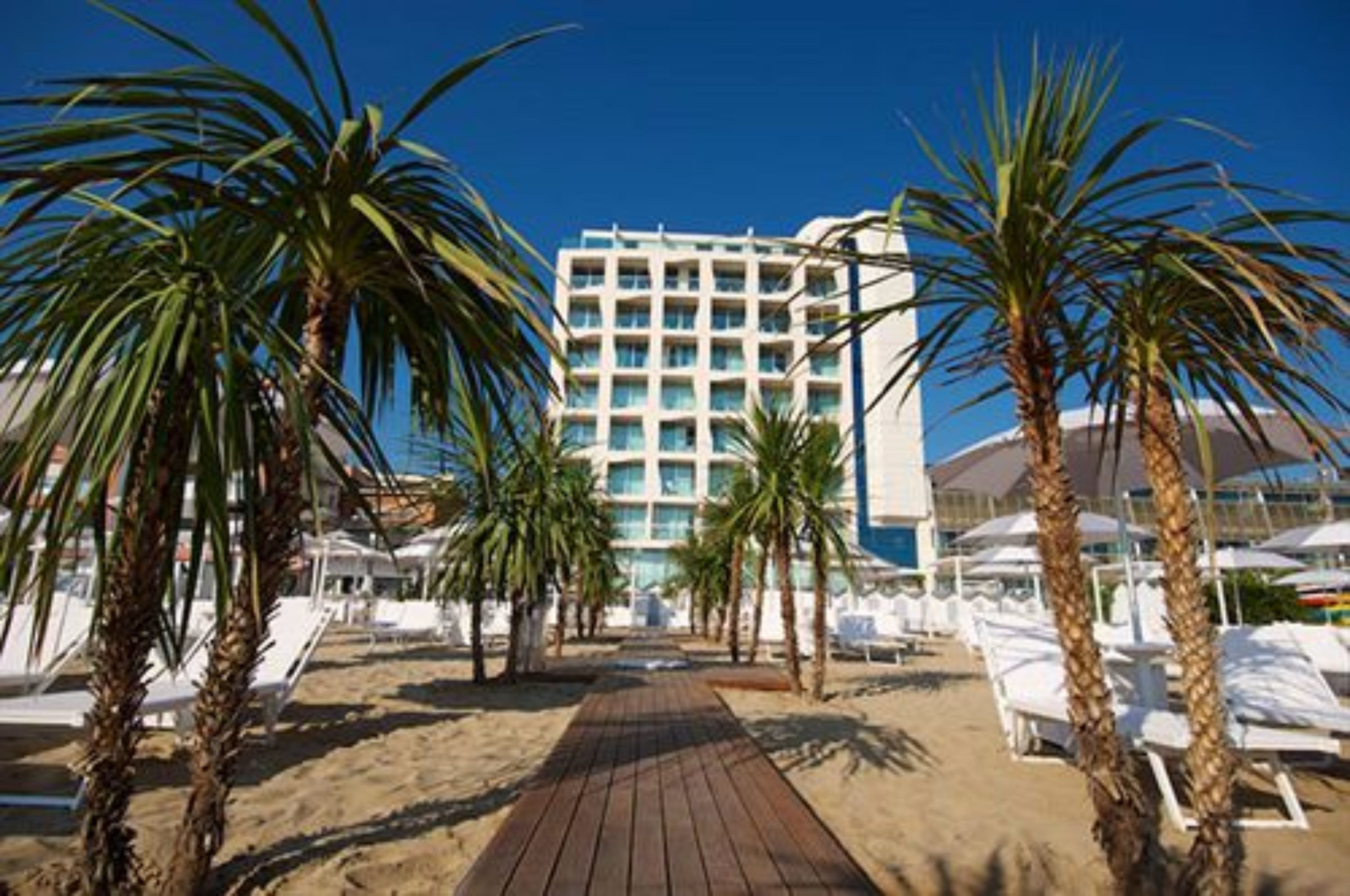 Un hotel frente al mar con un pasillo de madera que lleva a sillas reclinables blancas y palmeras.