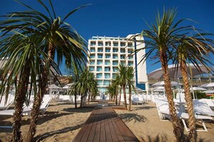 Un hotel frente al mar con un pasillo de madera que lleva a sillas reclinables blancas y palmeras.