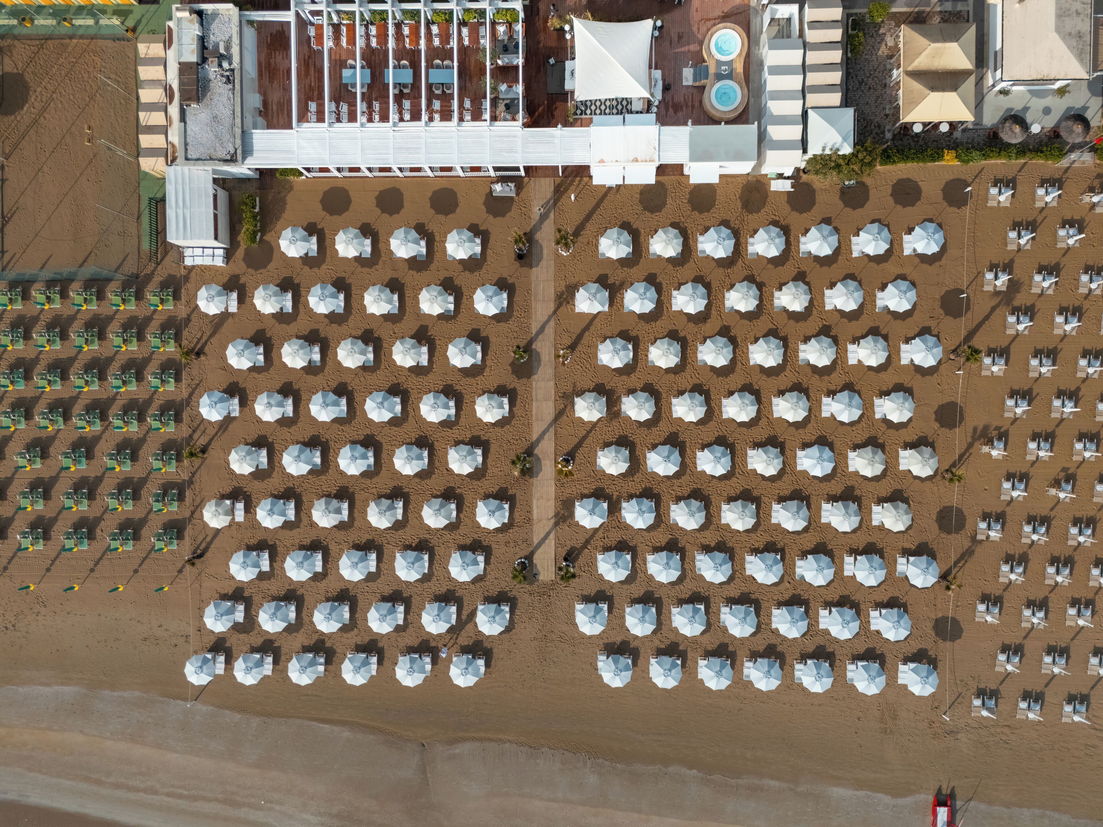 Vista aérea de un complejo turístico en la playa con hileras de sombrillas y sillas, un área de piscina y un edificio con fachada blanca.