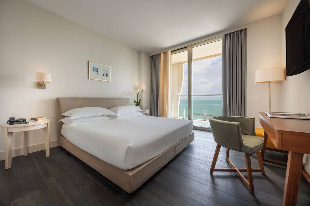 A hotel room with a large bed, a desk, a chair, a TV, and a view of the ocean.