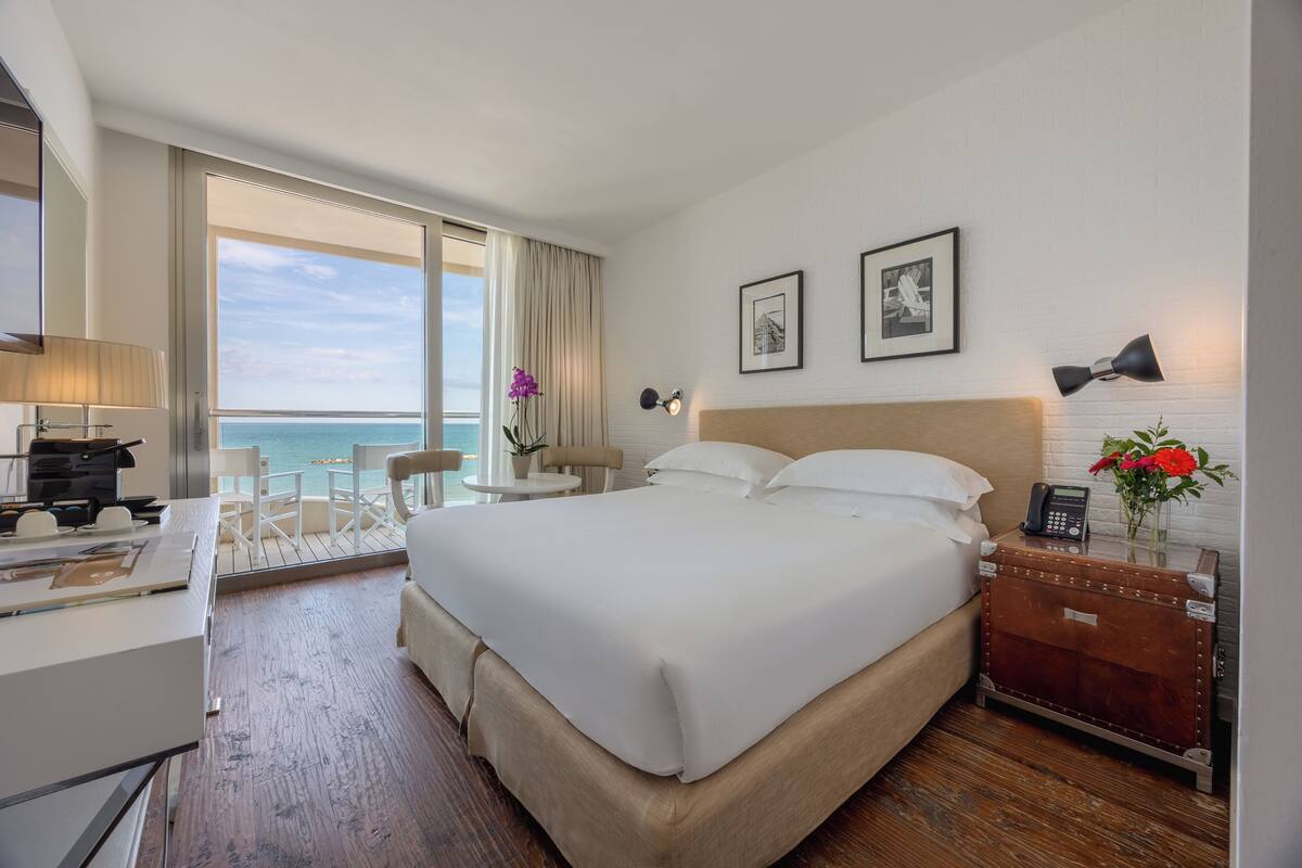 A hotel room with a large bed, a desk, a chair, a small table with a lamp, and a view of the ocean.