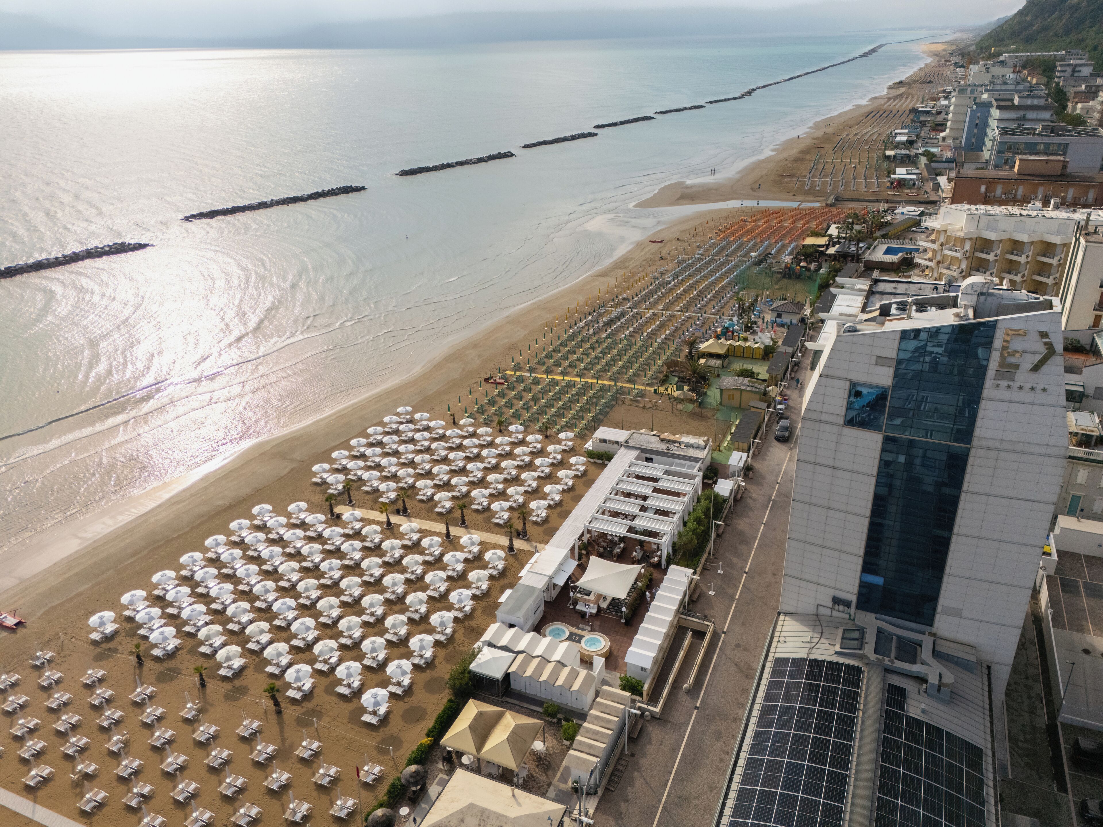 An aerial view of a beachside hotel with rows of sunbeds and umbrellas, a swimming pool, and a modern building with a glass facade.