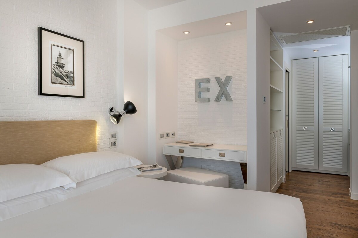 A modern hotel room with a large bed, a bedside table, a desk with a chair, a wardrobe, and a wall-mounted light fixture.