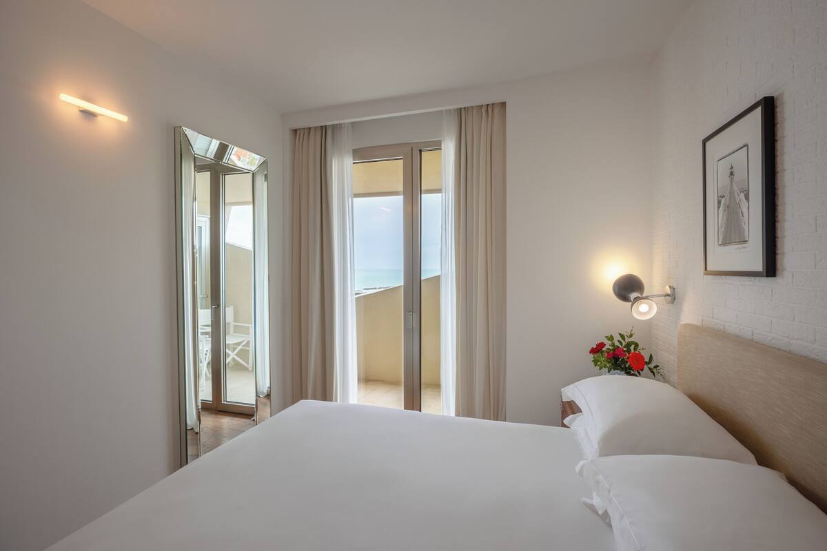 A hotel room with a bed, a nightstand with a lamp, a framed picture on the wall, and a view of the balcony and outside.