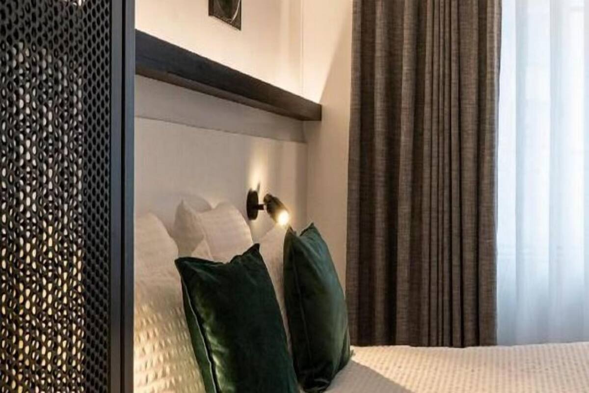 Une chambre d’hôtel avec un lit, des oreillers vert foncé, une tête de lit dotée d’un luminaire et une fenêtre avec des rideaux.