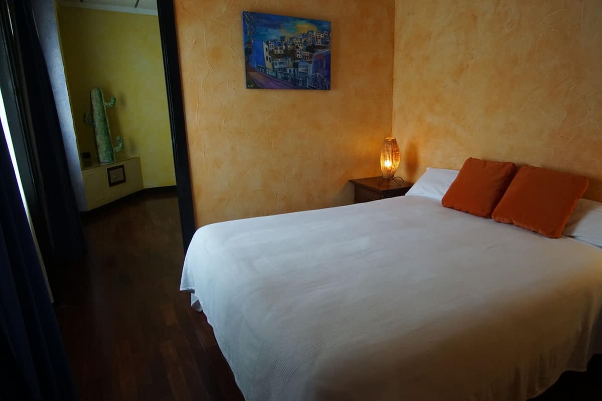 A neatly made bed with white linens and two orange pillows, a bedside lamp, and a painting of a coastal town on the wall.