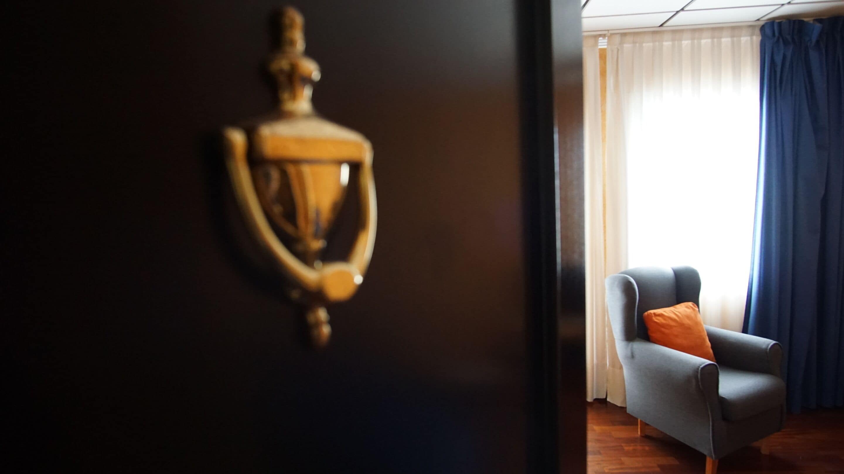 Een kamer met een donkerhouten deur, voorzien van een koperen deurbel, een grijze fauteuil met een oranje kussen en een raam met dunne witte gordijnen en een blauwe omslagdoek.