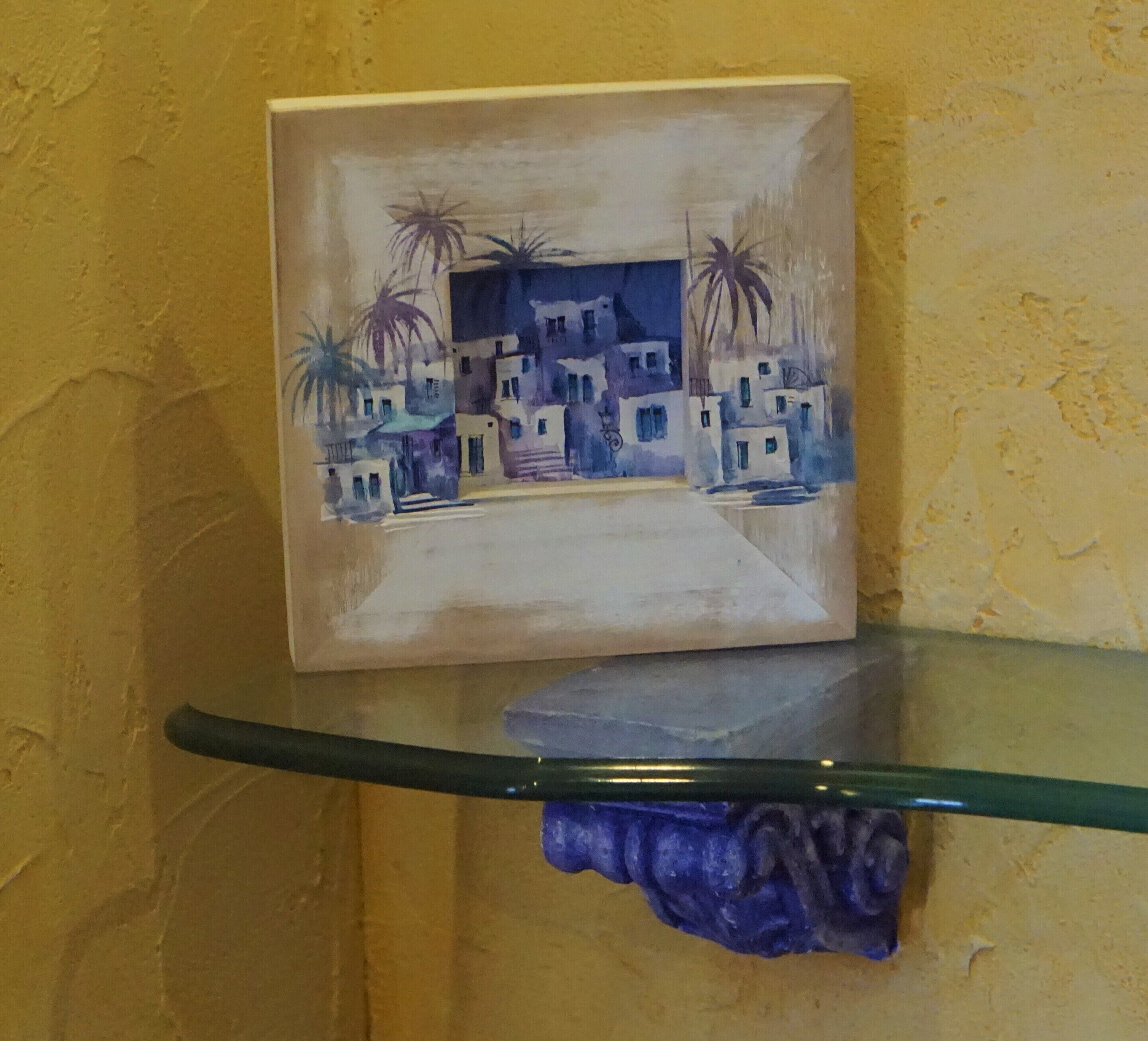 Un cuadro enmarco de un pueblo costero sobre una repisa de vidrio frente a una pared con textura.