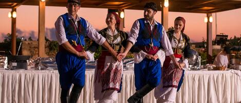 Un groupe de personnes, vêtues de costumes traditionnels, interprète une danse lors d’un événement en plein air, sur une scène couverte, avec des tables disposées en arrière-plan.