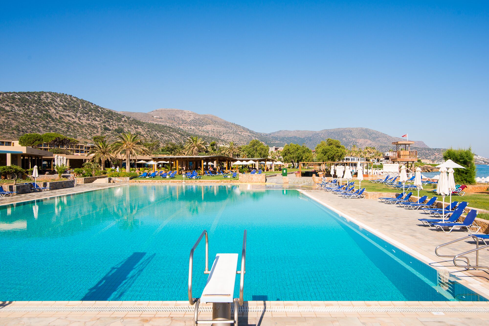 Ein großer Swimmingpool mit klarem, blauem Wasser, umgeben von Liegestühlen und Sonnenschirmen, vor einer Bergkulisse und wolkenlosem Himmel.