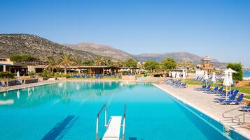 Ein großer Swimmingpool mit klarem, blauem Wasser, umgeben von Liegestühlen und Sonnenschirmen, vor einer Bergkulisse und wolkenlosem Himmel.