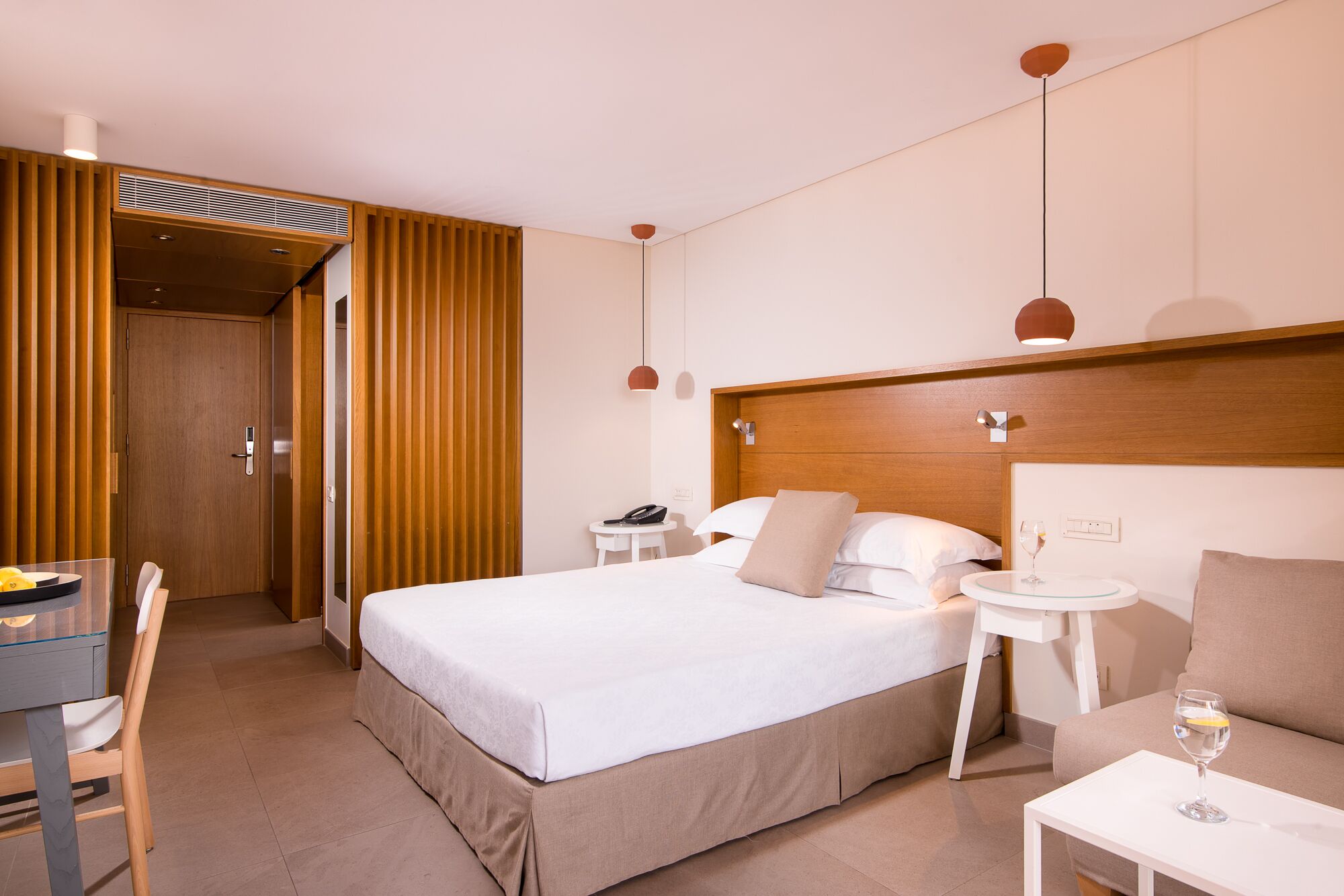 A hotel room with a bed, bedside table, chair, and a small desk with a lamp.