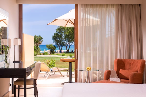A hotel room with a bed, a desk, a chair, and a table with a glass top. There is a view of the beach and ocean through the window.