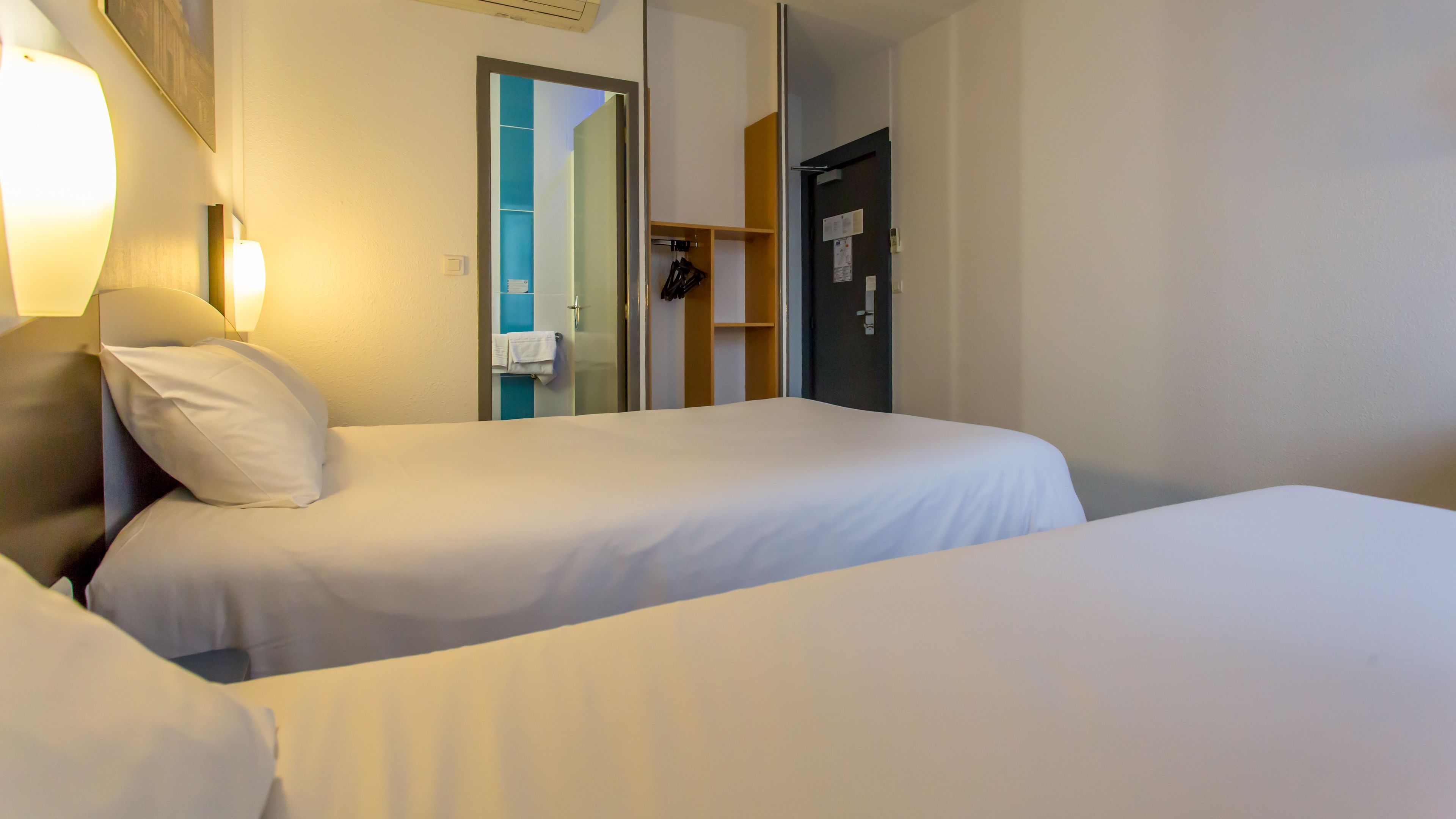 Ett hotellrum med två separata sängar, ett nattduksbord, en lampa, en spegel och en dörr som leder till ett badrum.