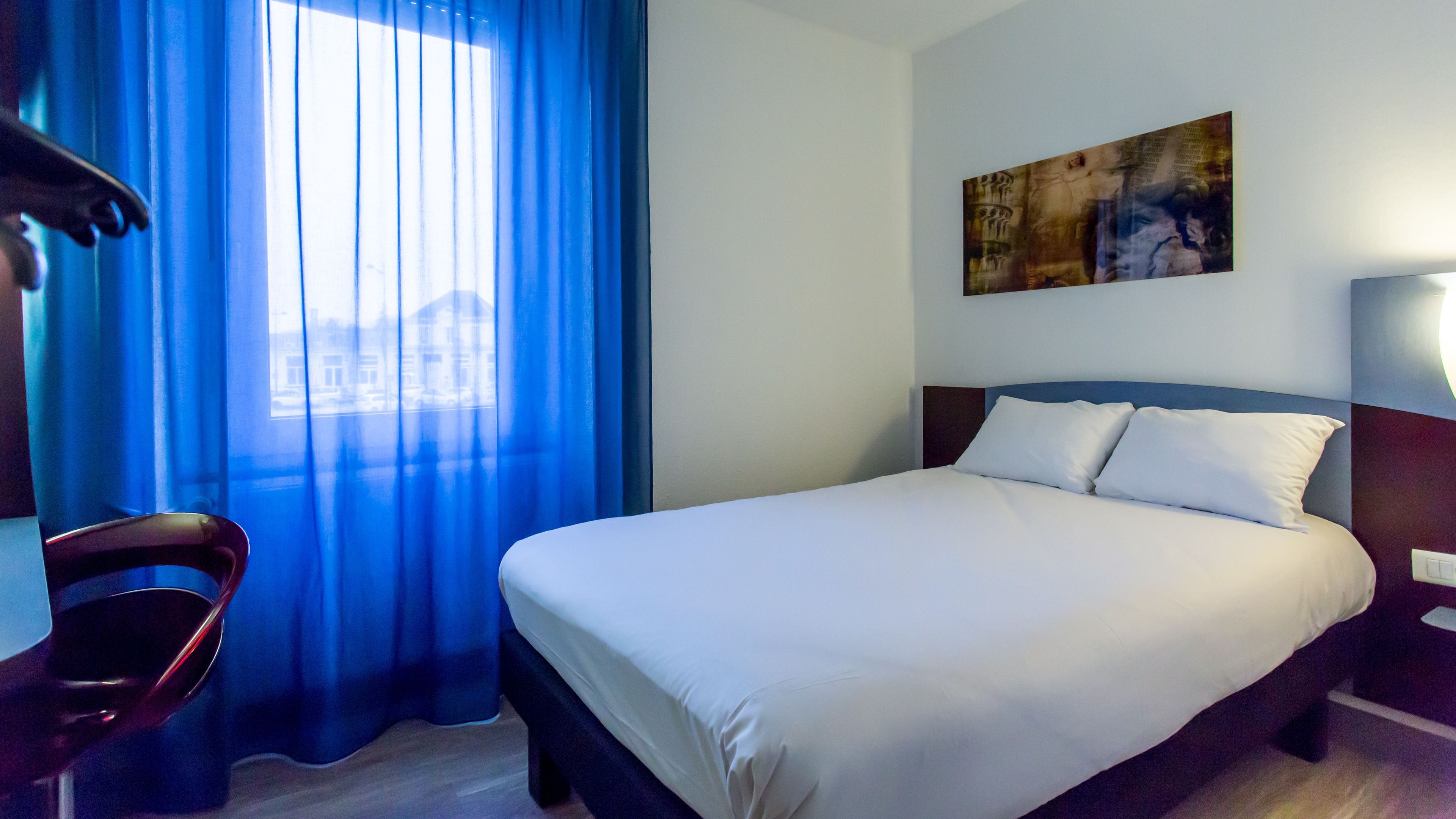 Habitación de hotel con cama, una silla, un cuadro en la pared y ventana con cortinas.