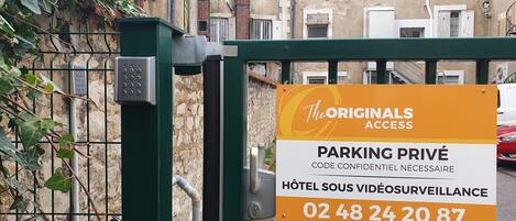 Une entrée fermée par un portail, agrémentée d’un panneau indiquant qu’il s’agit d’un parking privé, avec un numéro de téléphone à contacter et un système de surveillance.