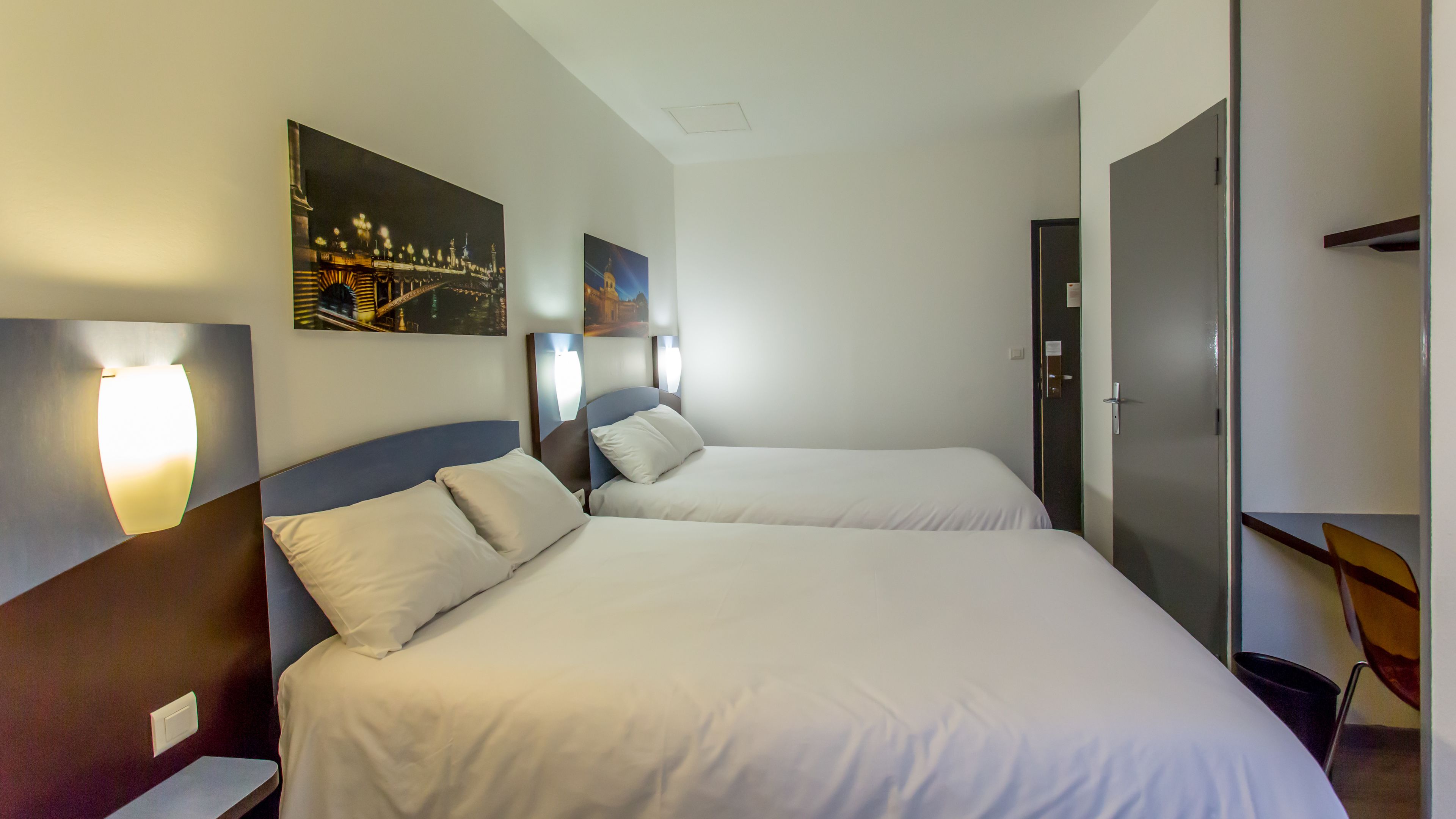 A hotel room with two beds, a nightstand, a wall-mounted light, and a door.