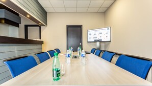 Een congreszaal met een lange tafel, blauwe stoelen, waterflesjes en een presentatiescherm.