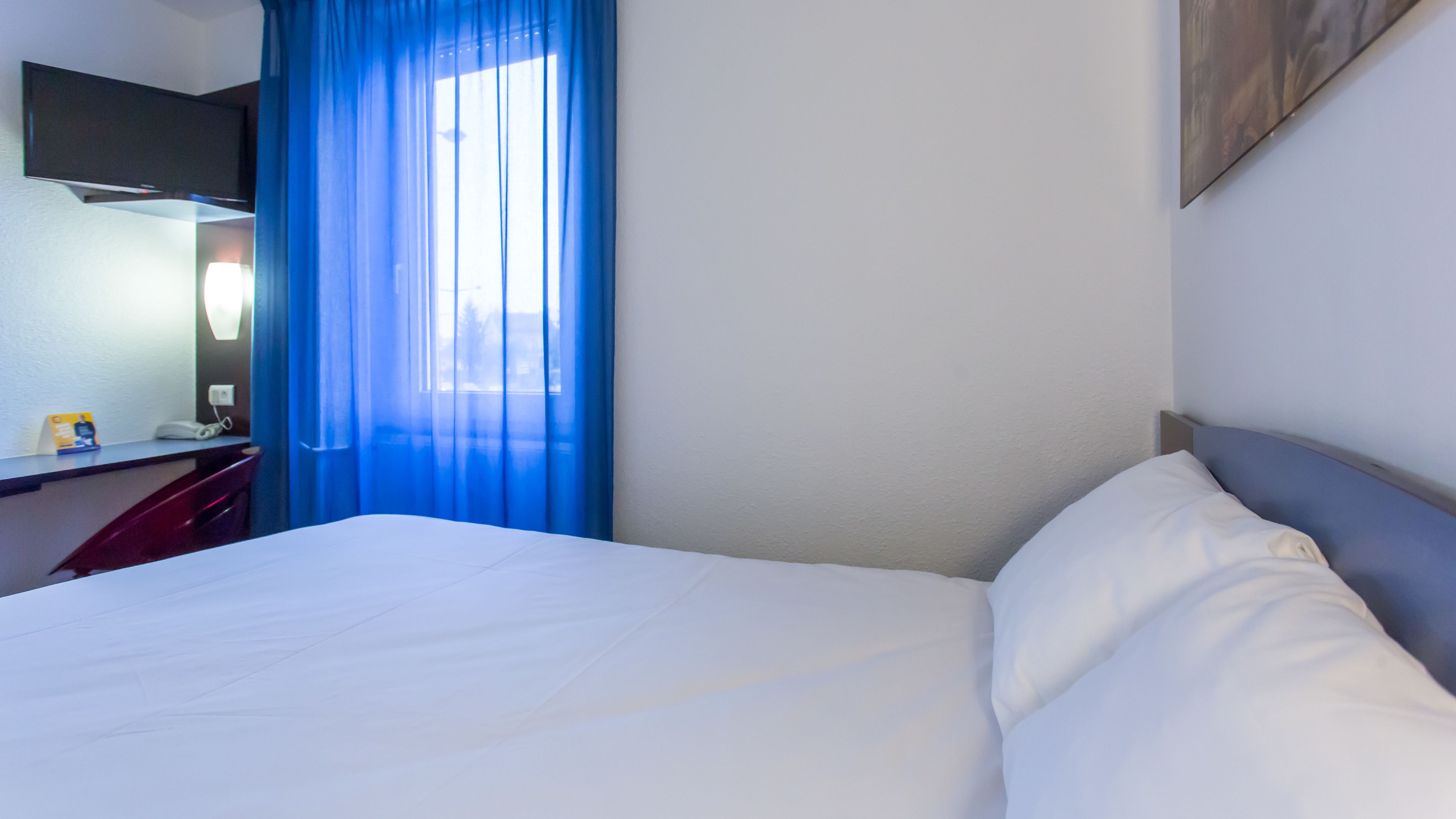 A hotel room with a bed, a desk, a flat-screen TV, and a window with blue curtains.