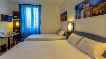 A hotel room with two beds, a desk, a chair, a TV, and a large window with blue curtains.