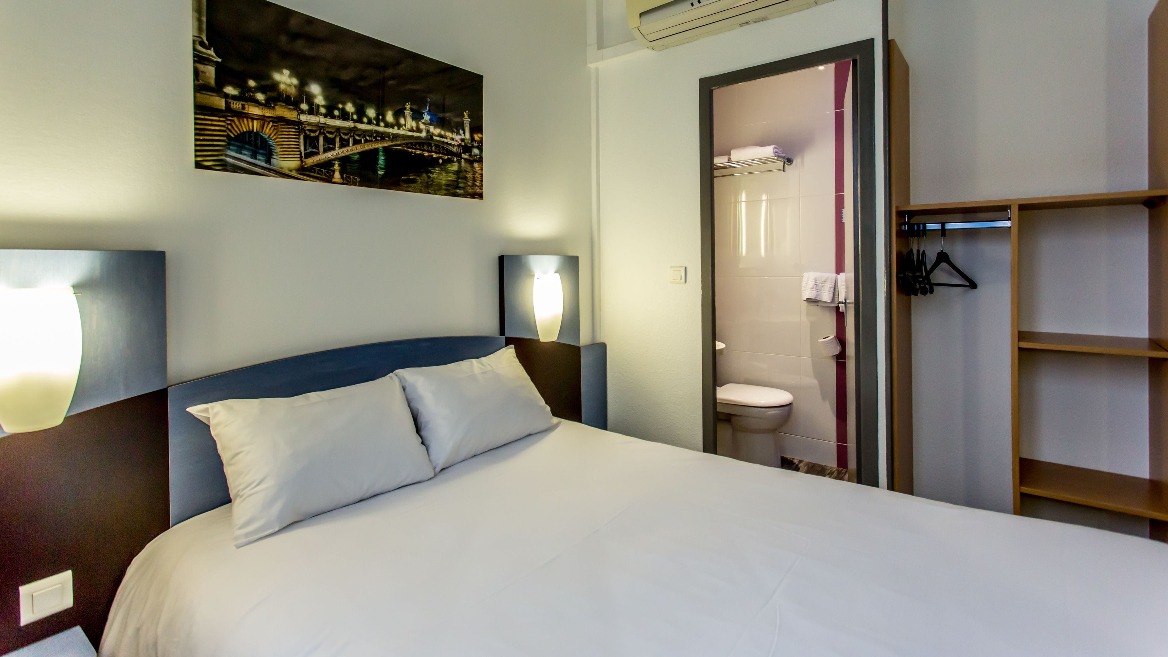 Una habitación de hotel con cama, mesita de noche, lámpara de pared, baño con inodoro y estante con perchas.