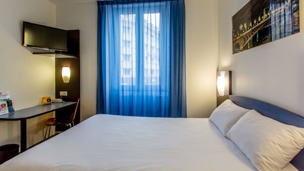 Een hotelkamer met een bed, een bureau, een stoel, een televisie en een raam met blauwe gordijnen.