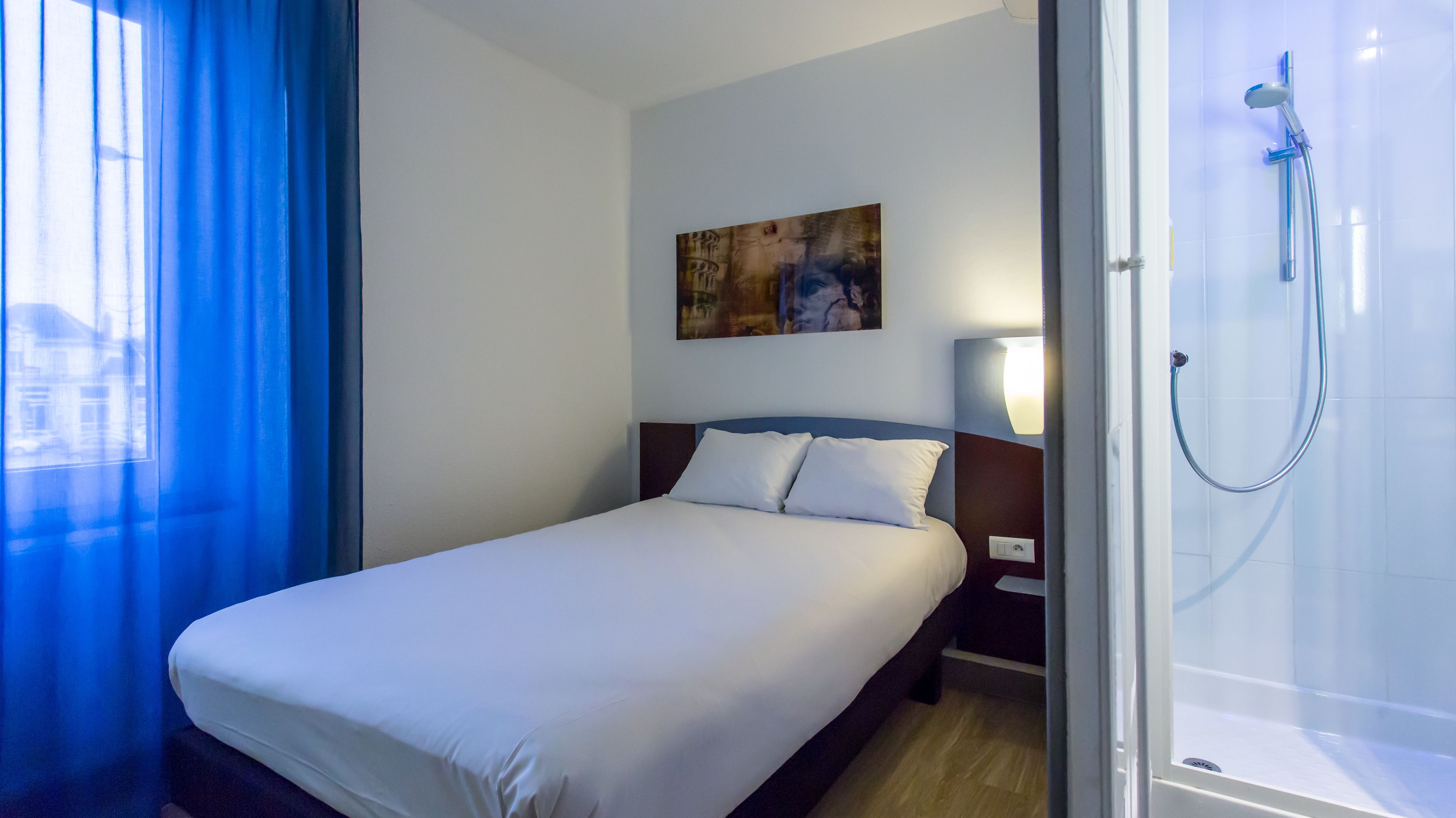 A hotel room with a bed, a painting on the wall, a nightstand, a lamp, and a shower.