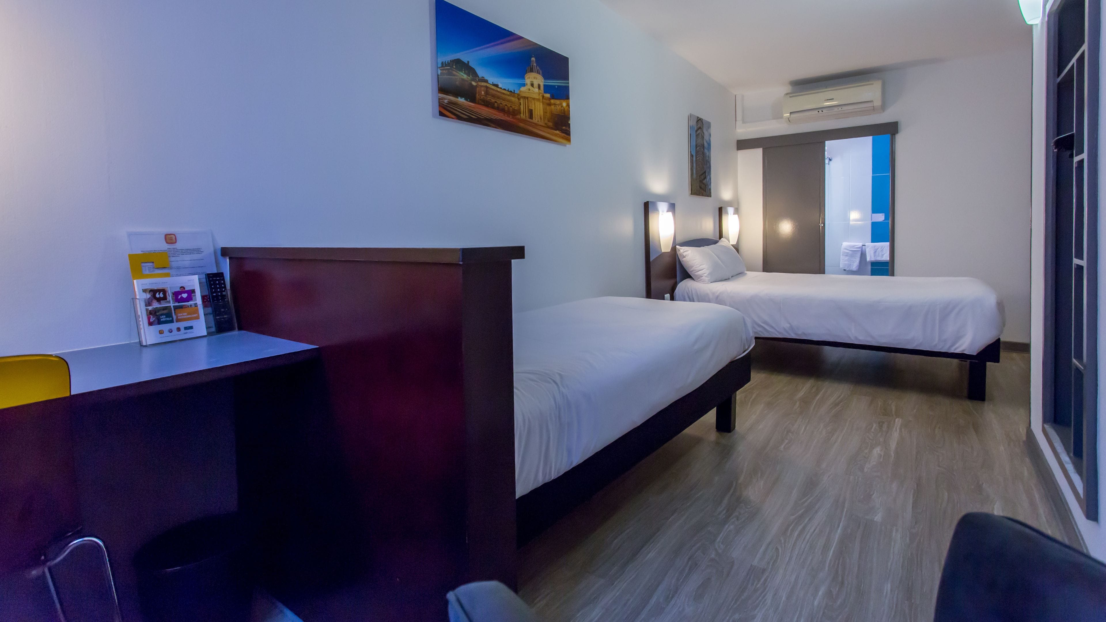 Ein Hotelzimmer mit zwei Einzelbetten, einem Schreibtisch mit Computer und einem Fenster mit Blick auf ein Gebäude.