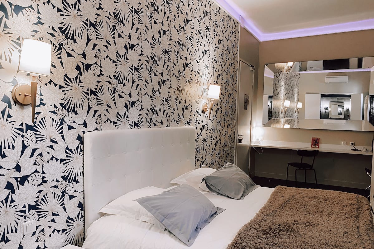 Habitación de hotel con cama, mesita de noche, espejo y baño visible a través de una puerta abierta.