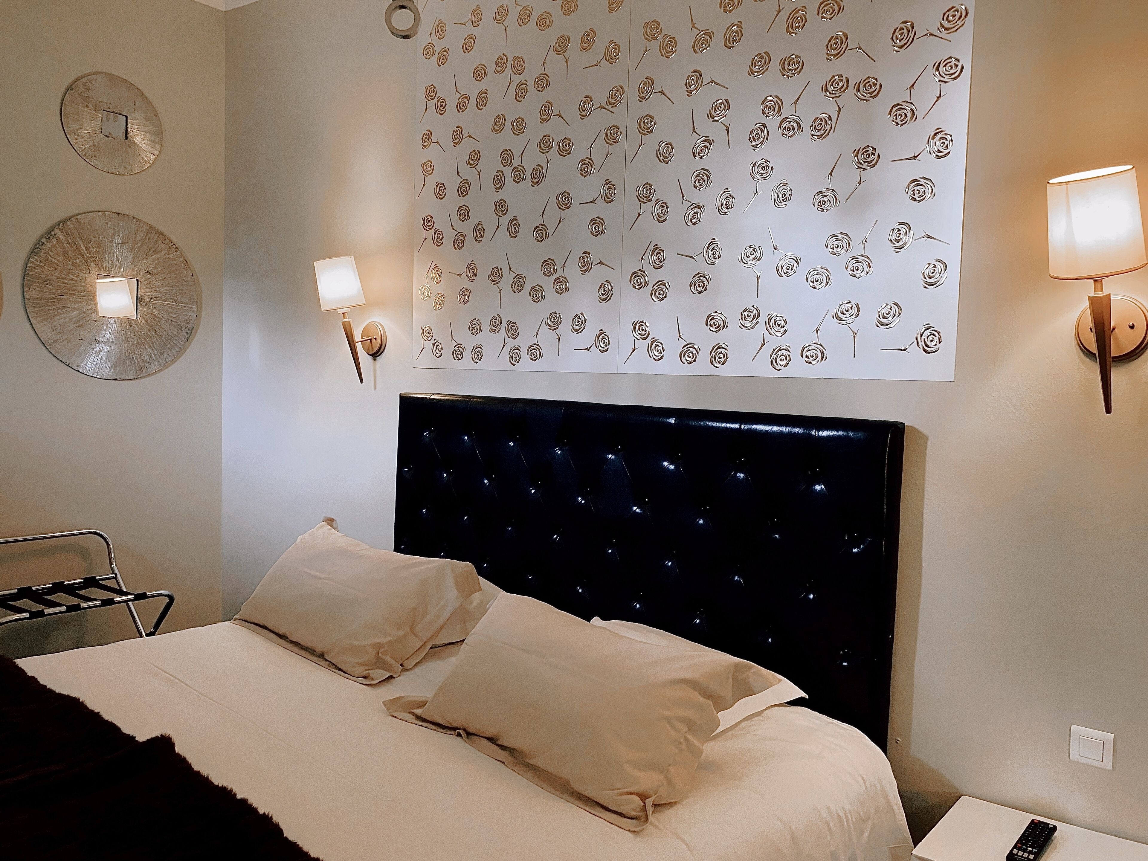 A neatly made bed with two pillows, a wall clock, a decorative tapestry with a floral pattern, and two wall-mounted lamps.