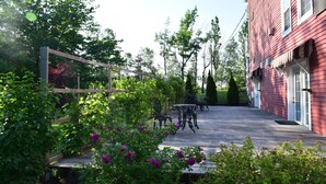 Una terraza de madera con mobiliario exterior, rodeada de exuberante vegetación y un edificio rojo al fondo.