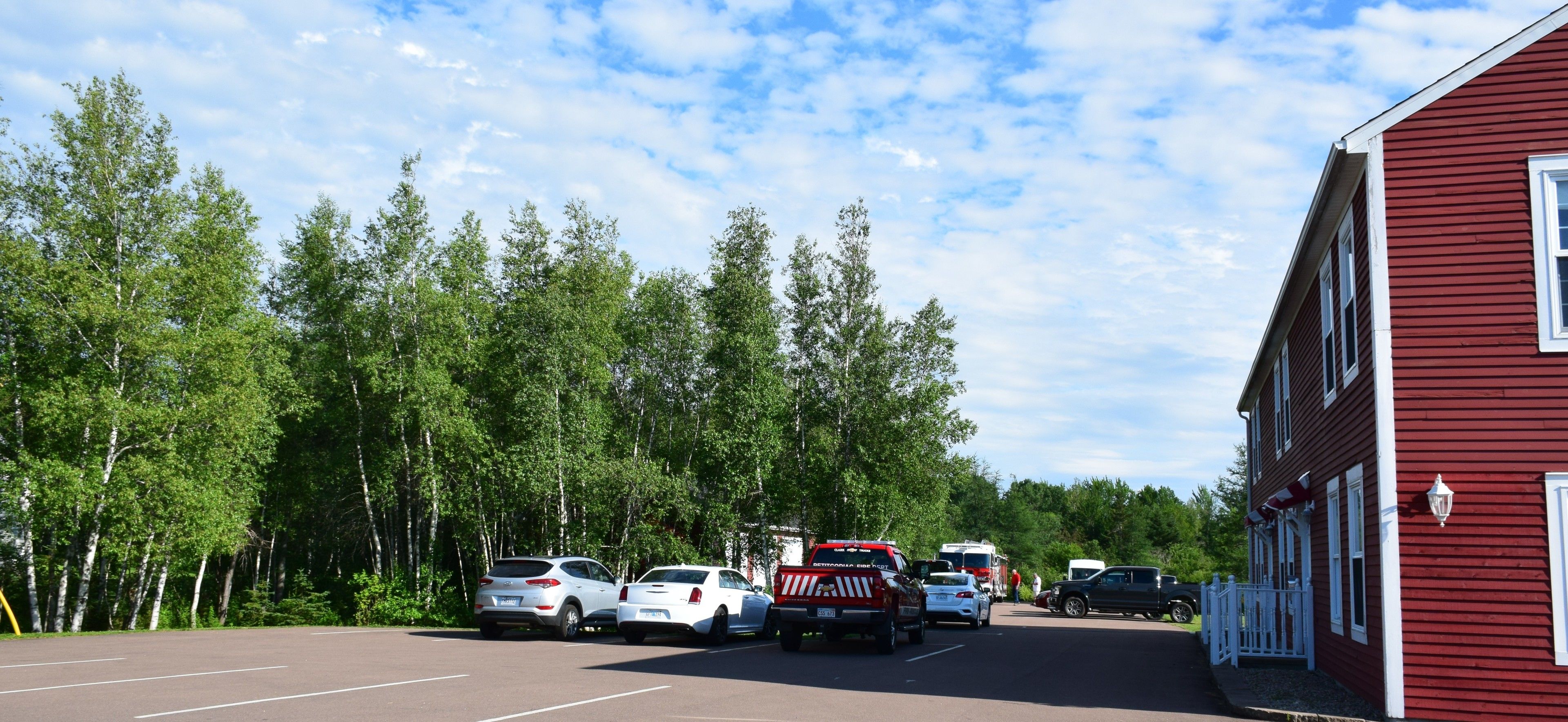 A parking lot with several cars and emergency vehicles, surrounded by trees and a red building.