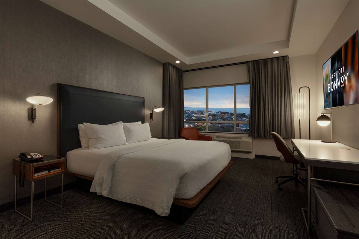 Ein Hotelzimmer mit einem großen Bett, einem Schreibtisch, einem Sessel und Blick auf die Stadt durch ein großes Fenster.